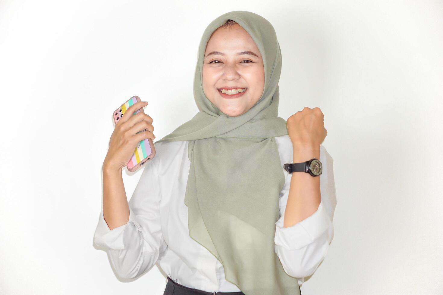 A cheerful asian woman in a green hijab celebrates a victory while holding her phone and smiling. photo
