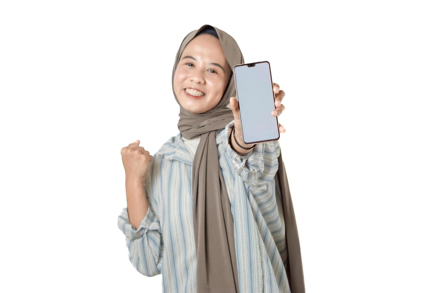 A cheerful hijab-wearing woman clenches her hands into fists while holding a smartphone, expressing happiness photo