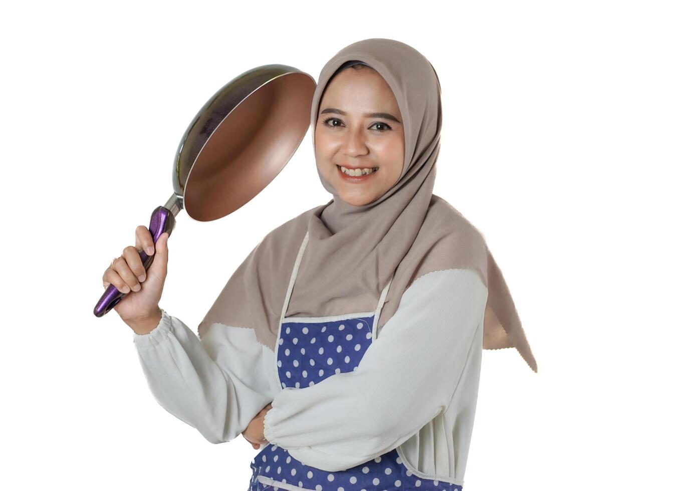A cheerful woman in a hijab holding a frying pan and wearing a polka dot apron, ready to cook with a smile, isolated on a white background. photo