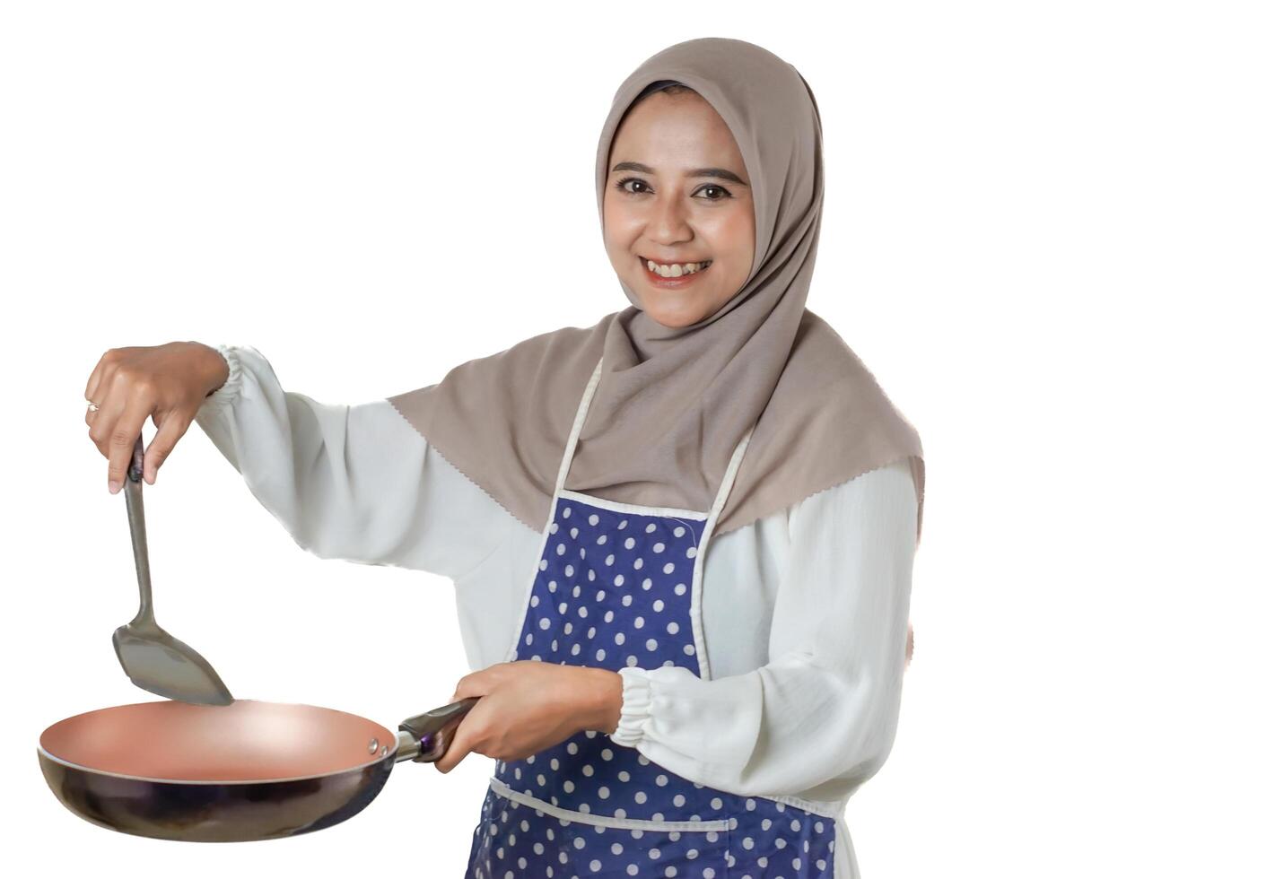 A woman in a hijab and a polka dot apron joyfully cooks with a frying pan, embodying the delight and warmth of home-cooked meals. photo