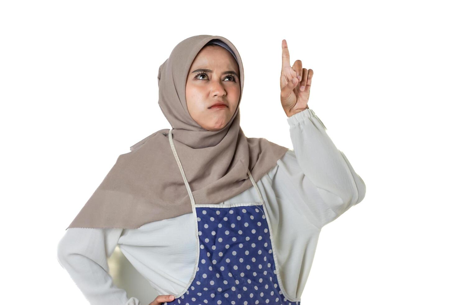 A woman wearing a hijab and a polka-dot apron points upwards with a thoughtful expression, contemplating something important. photo
