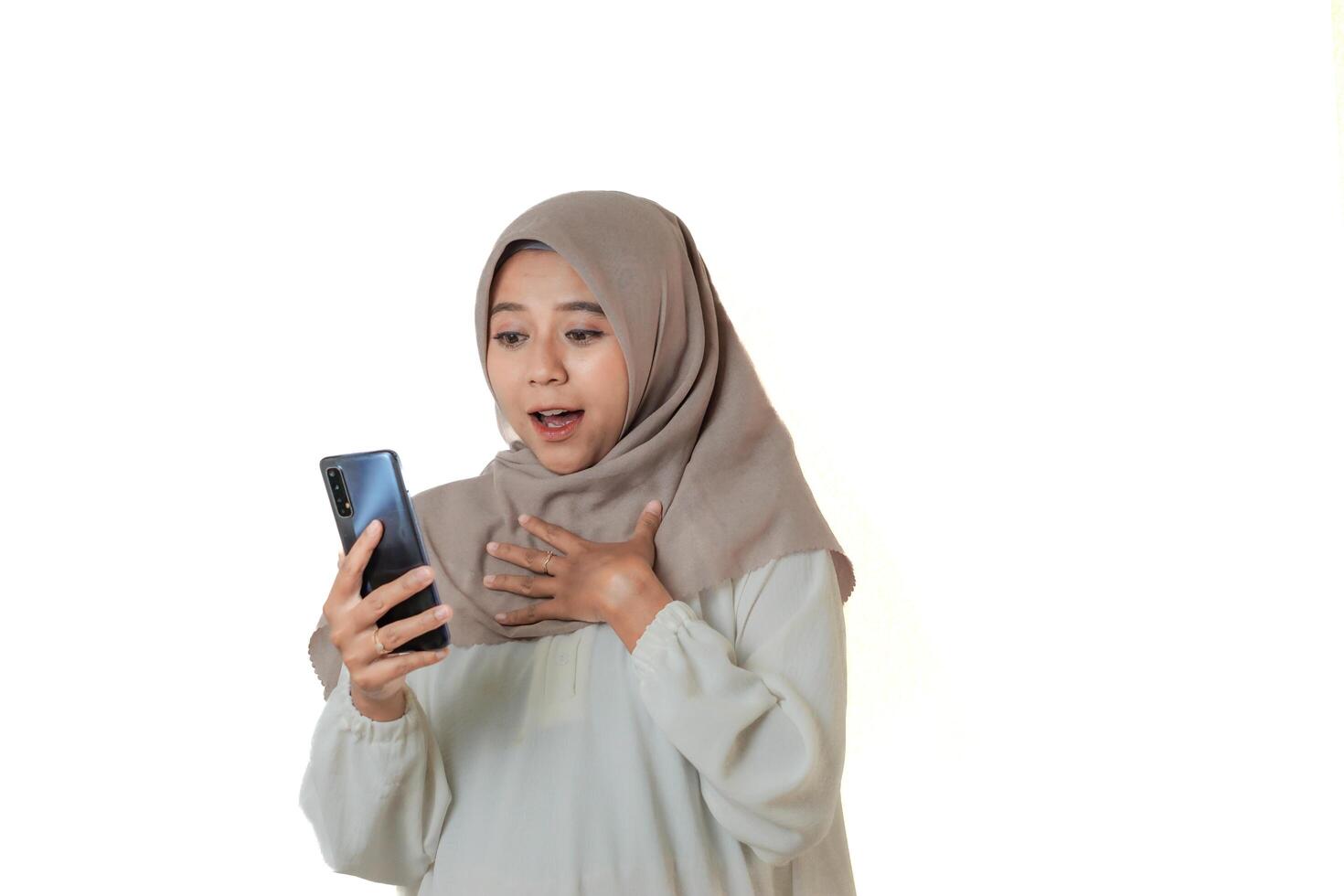 A woman in a beige hijab and white blouse looks excited and surprised while checking her smartphone. photo