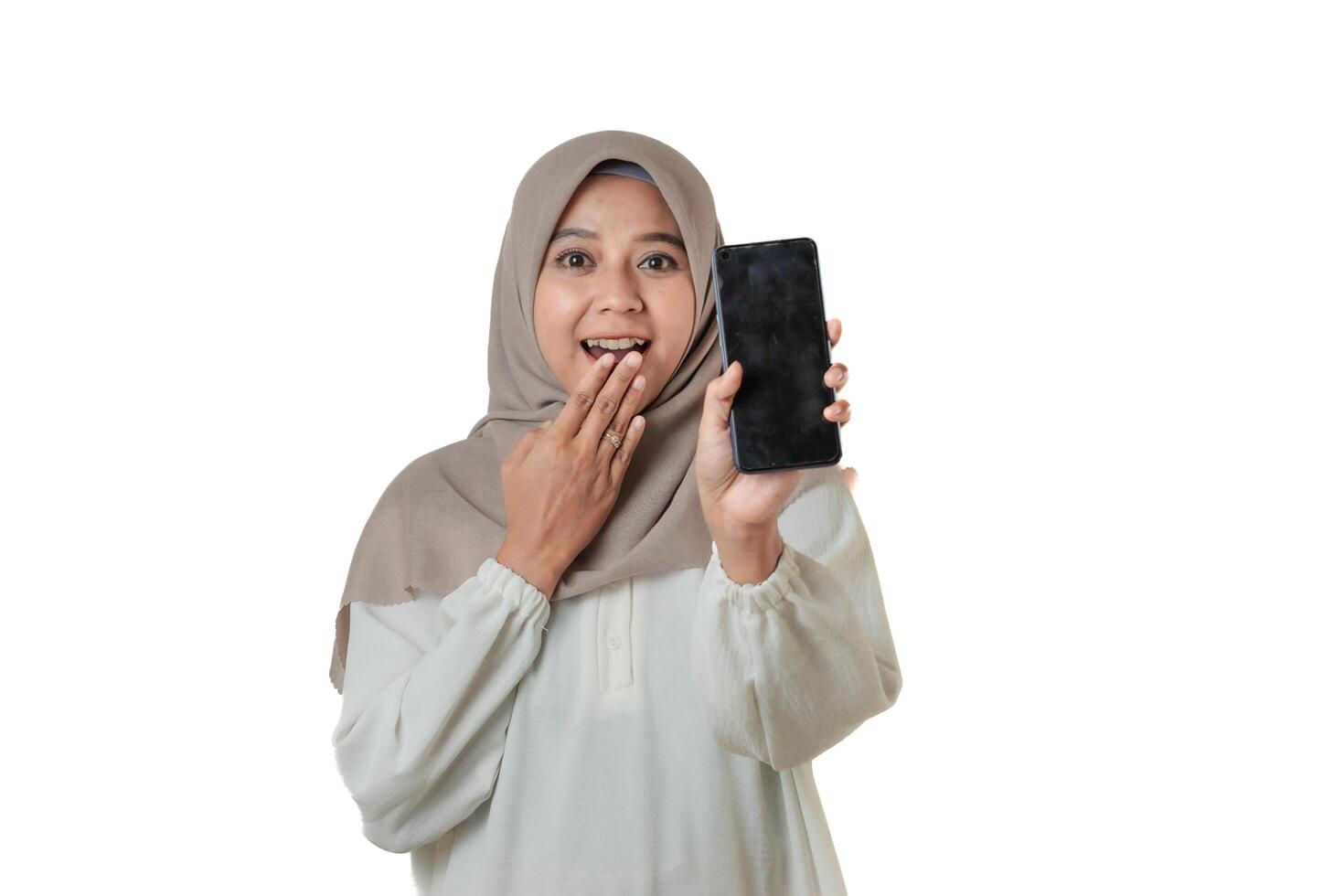 A woman in a beige hijab and white blouse, surprising, showing a smartphone. She holds her hand to her mouth in astonishment, expressing excitement. photo