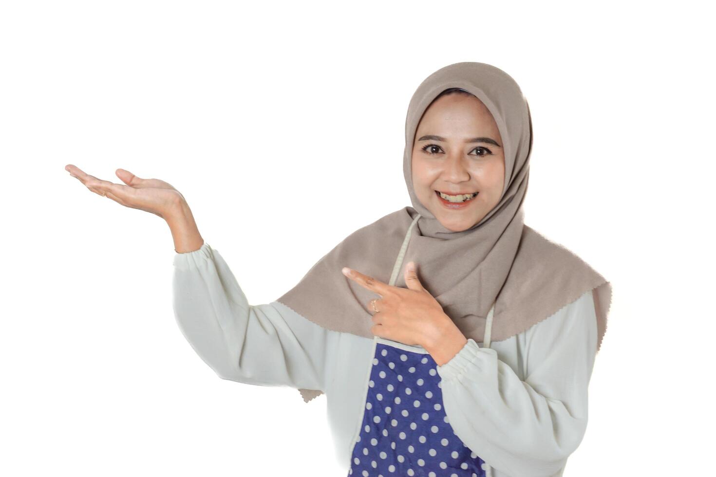A woman wearing a gray headscarf and polka-dot apron smiles while pointing and extending her hand, showing or exposing something. photo