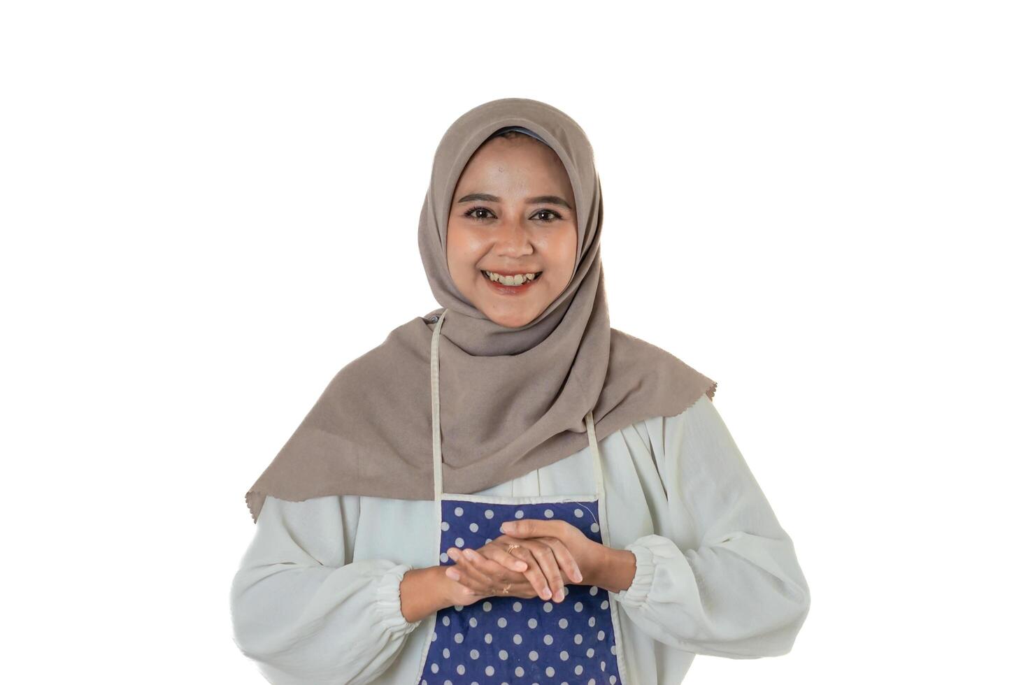 A cheerful woman wearing a beige hijab and a blue polka-dotted apron, smiling warmly at the camera. She exudes positivity and friendliness. photo