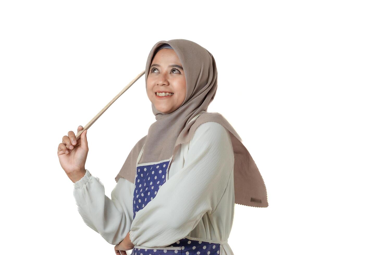 A woman wearing a hijab and a polka dot apron holds a kitchen utensil, smiling confidently, ready for cooking or baking in a bright, positive atmosphere. photo