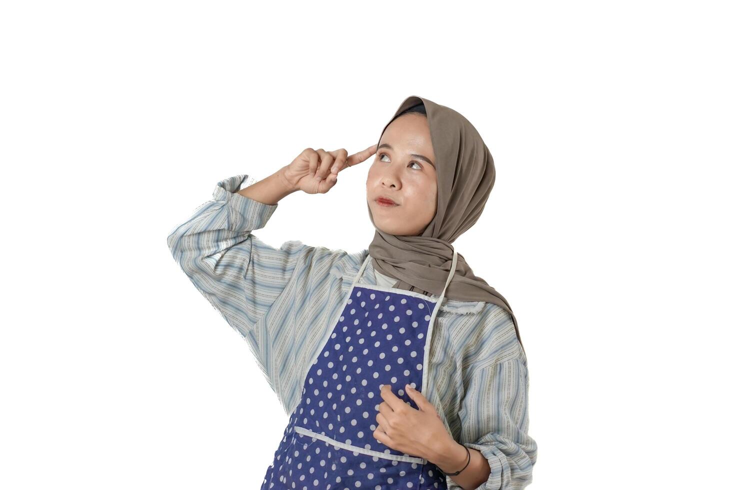 A woman in a striped shirt and polka dot apron, wearing a brown hijab, poses thoughtfully with her finger pointing to her forehead against a white background. photo