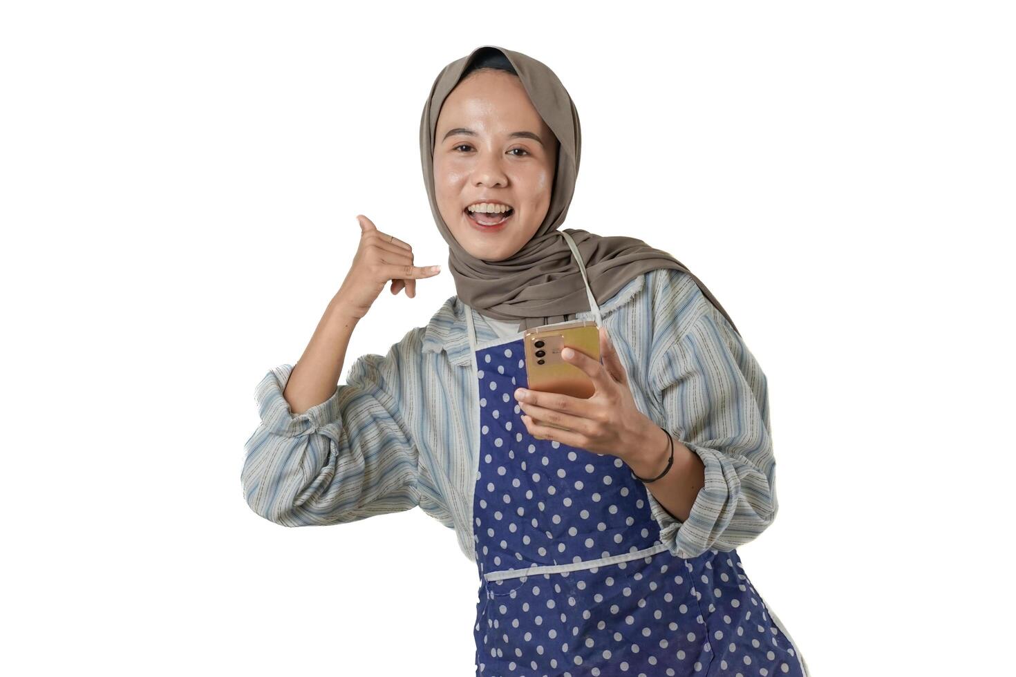 A woman wearing a hijab and a polka-dotted apron poses with a cheerful smile, making a call me hand gesture. She exudes warmth and friendliness. photo