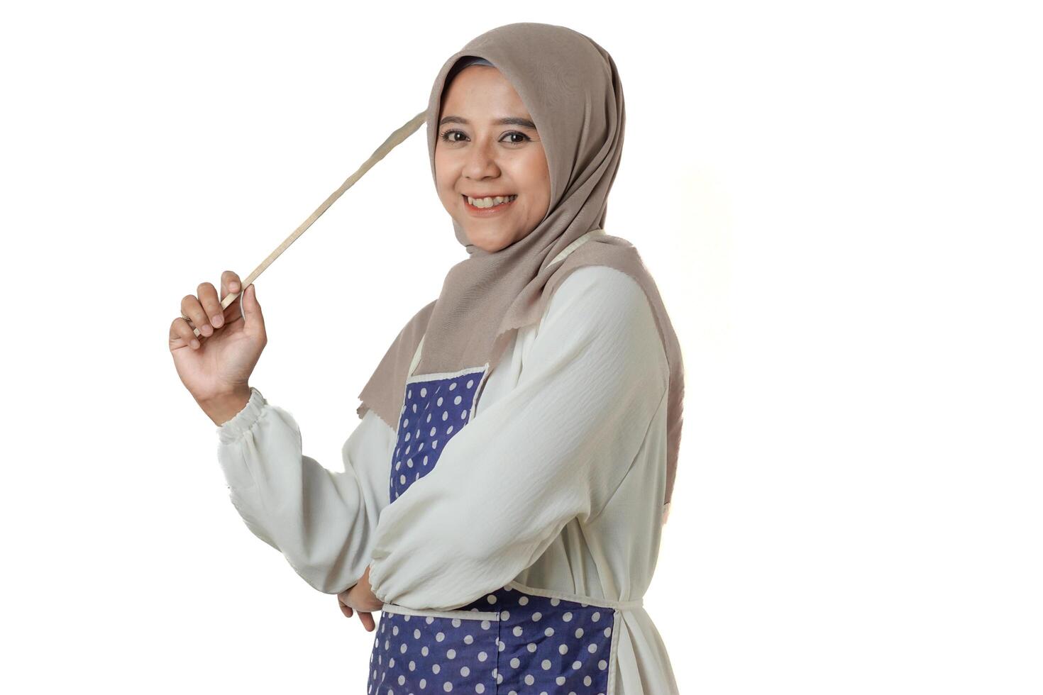 A woman wearing a hijab and a polka dot apron holds a kitchen utensil, smiling confidently, ready for cooking or baking in a bright, positive atmosphere. photo