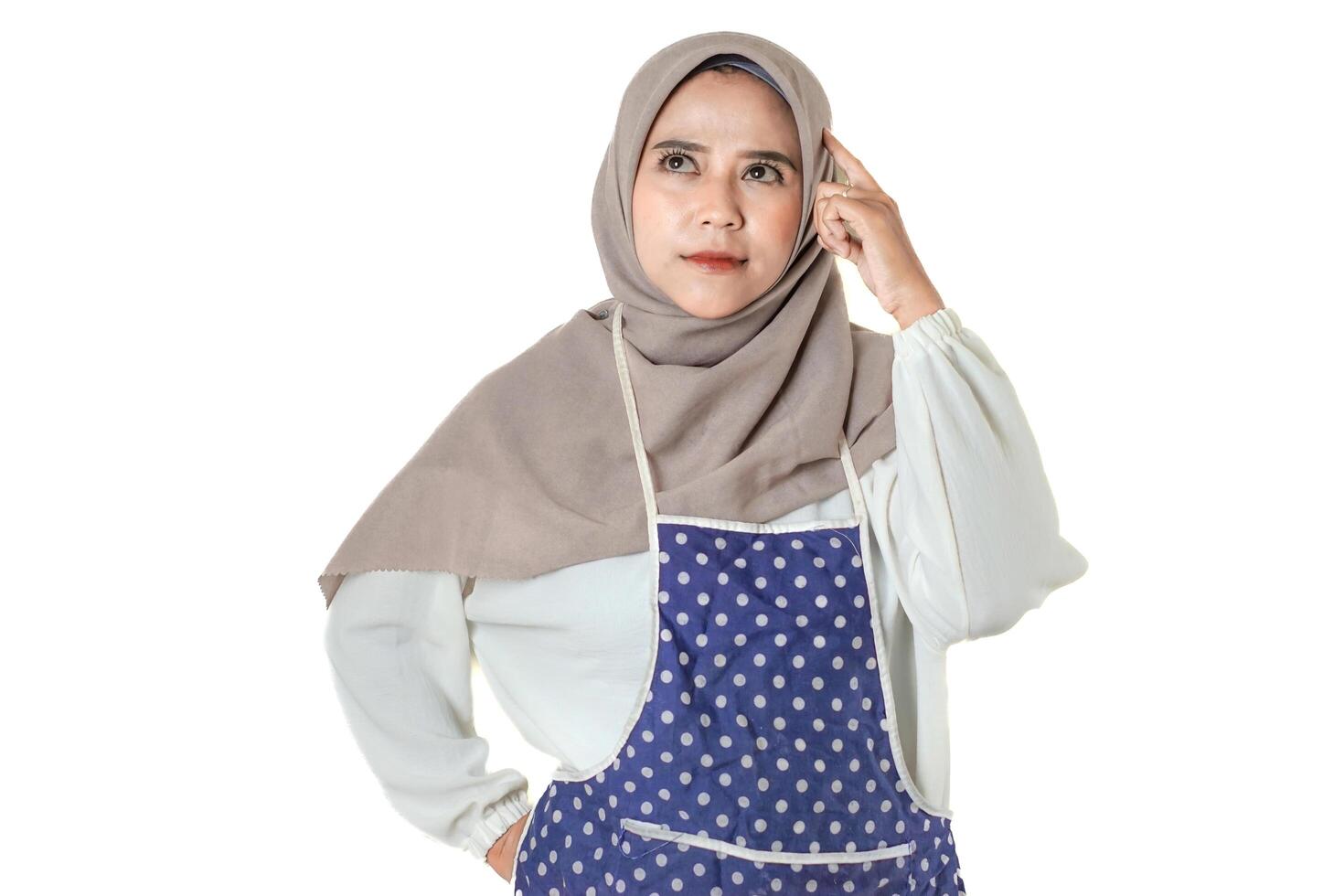 A woman in a hijab and apron is posing thoughtfully, with one hand touching her temple and the other on her hip, looking concerned or puzzled. photo