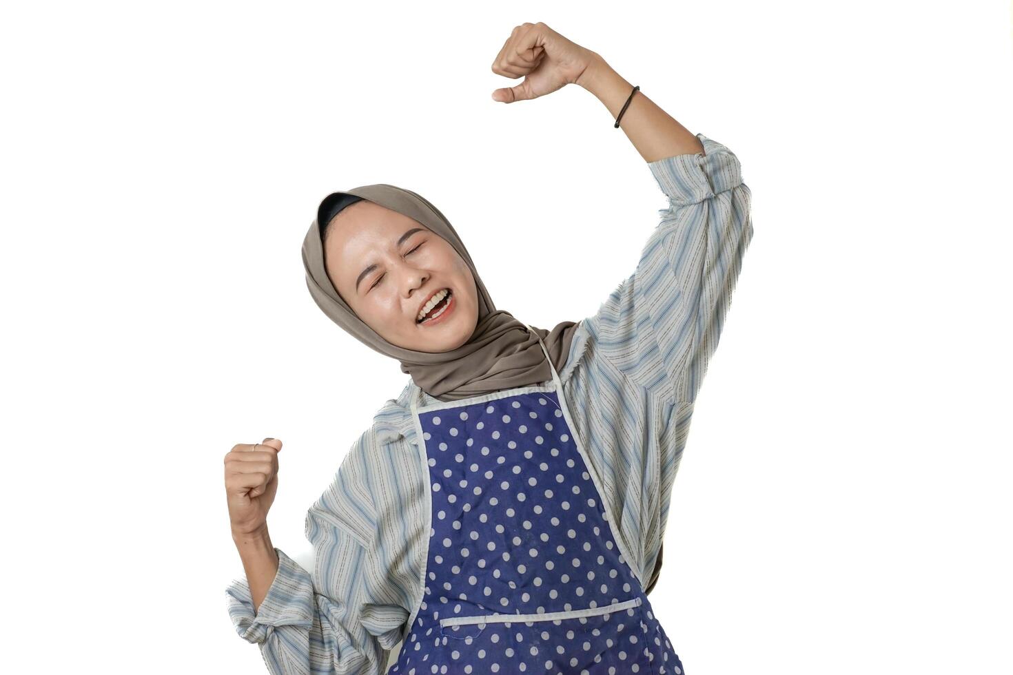 A joyful woman wearing a blue polka-dotted apron and a gray hijab is smiling and raising her arms in a celebratory gesture, expressing pride and happiness. photo