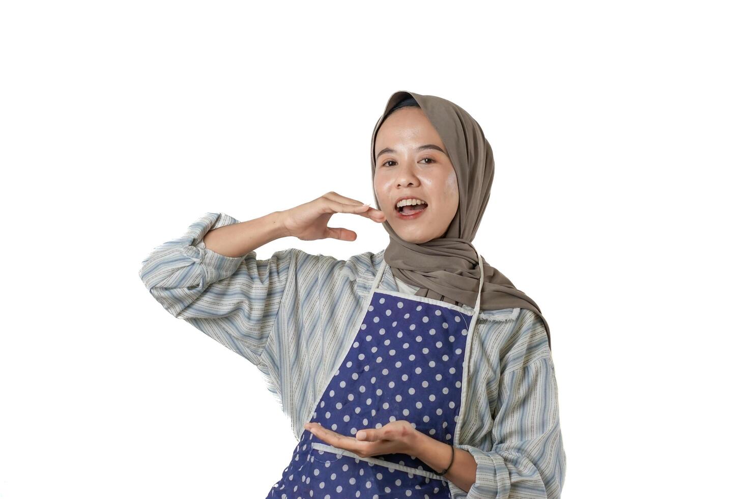 A woman wearing a hijab and an apron smiles and makes a hand gesture, possibly demonstrating something related to cooking or food presentation. photo