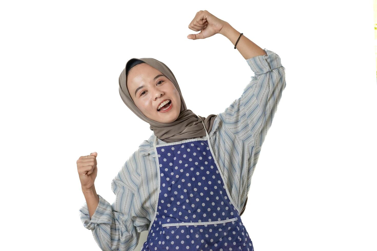 A joyful woman wearing a blue polka-dotted apron and a gray hijab is smiling and raising her arms in a celebratory gesture, expressing pride and happiness. photo