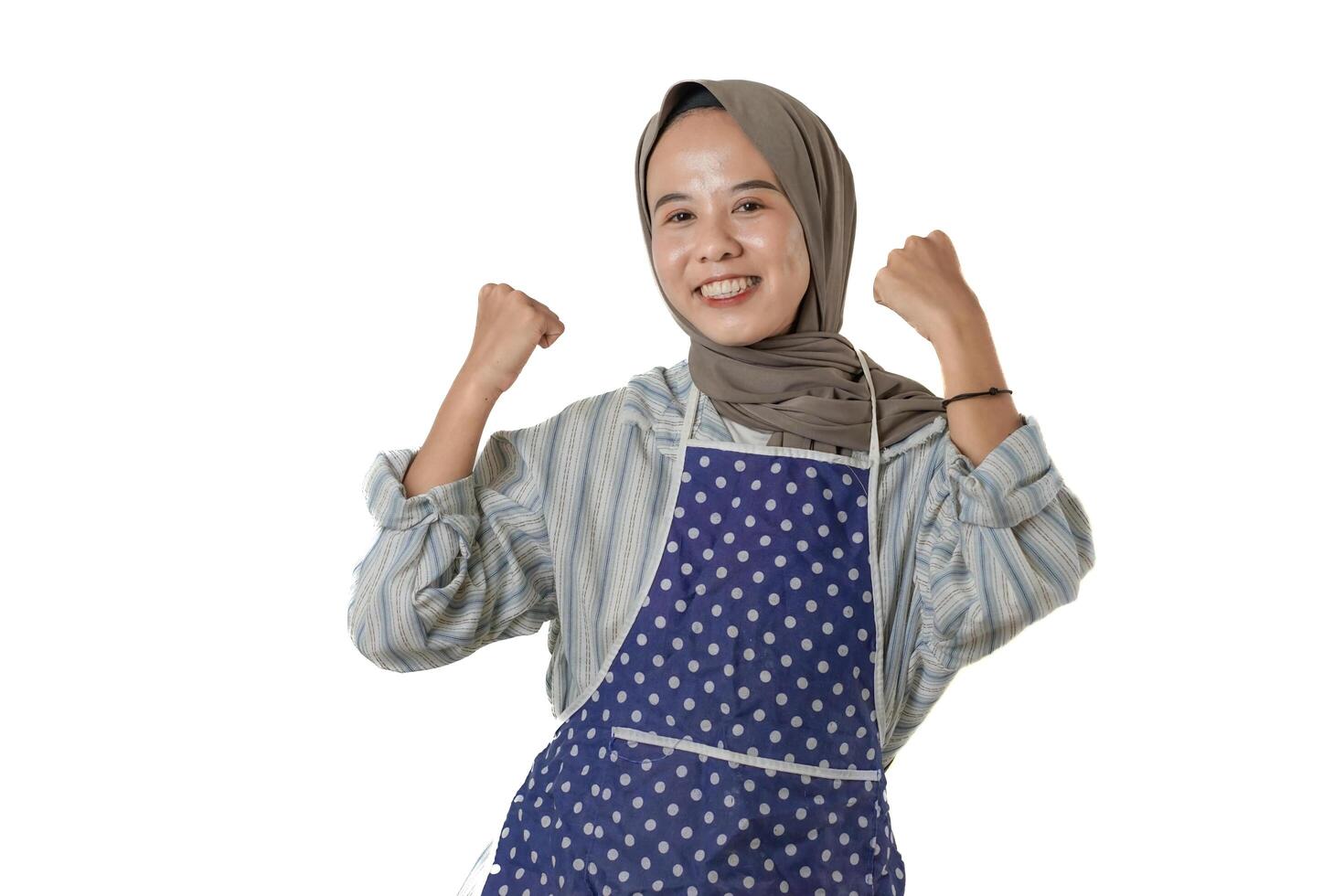 A joyful woman wearing a blue polka-dotted apron and a gray hijab is smiling and raising her arms in a celebratory gesture, expressing pride and happiness. photo