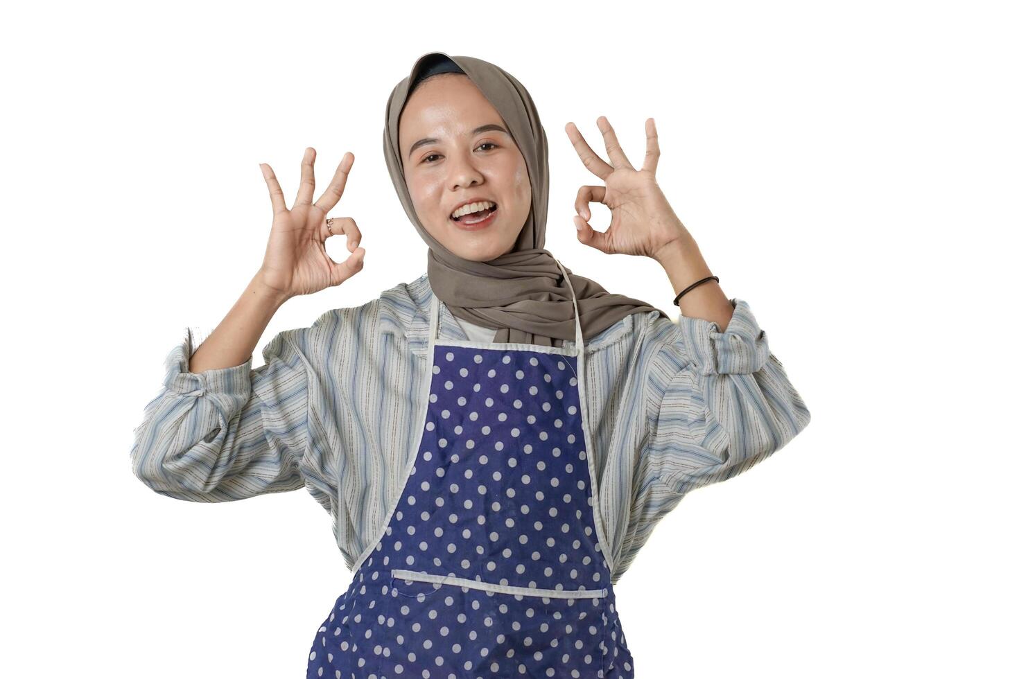 A cheerful woman wearing a blue polka-dot apron and a striped shirt, showing OK hand signs, expressing joy and satisfaction while preparing to cook. photo