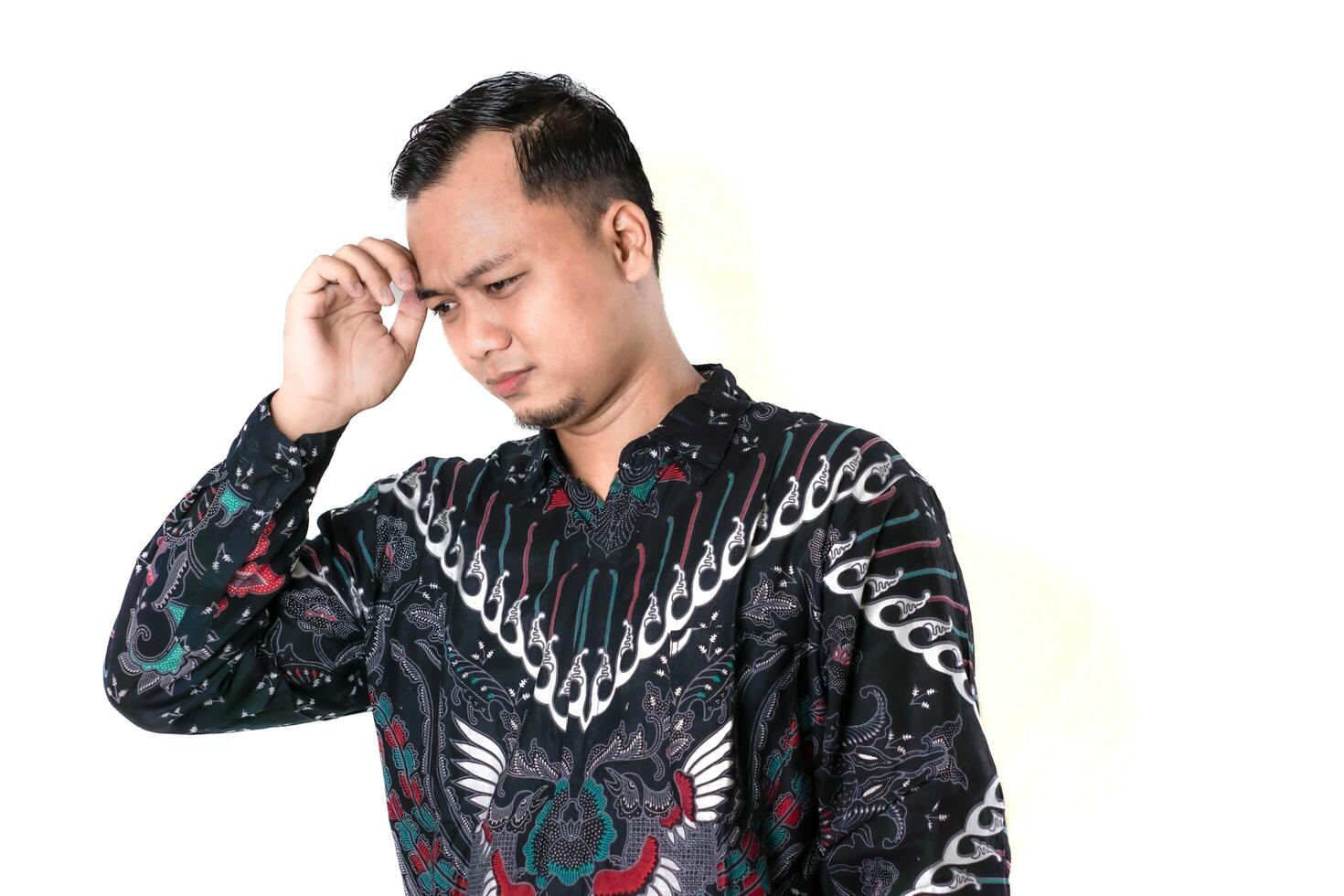 A man in a colorful batik shirt stands against a white background, deep in thought photo