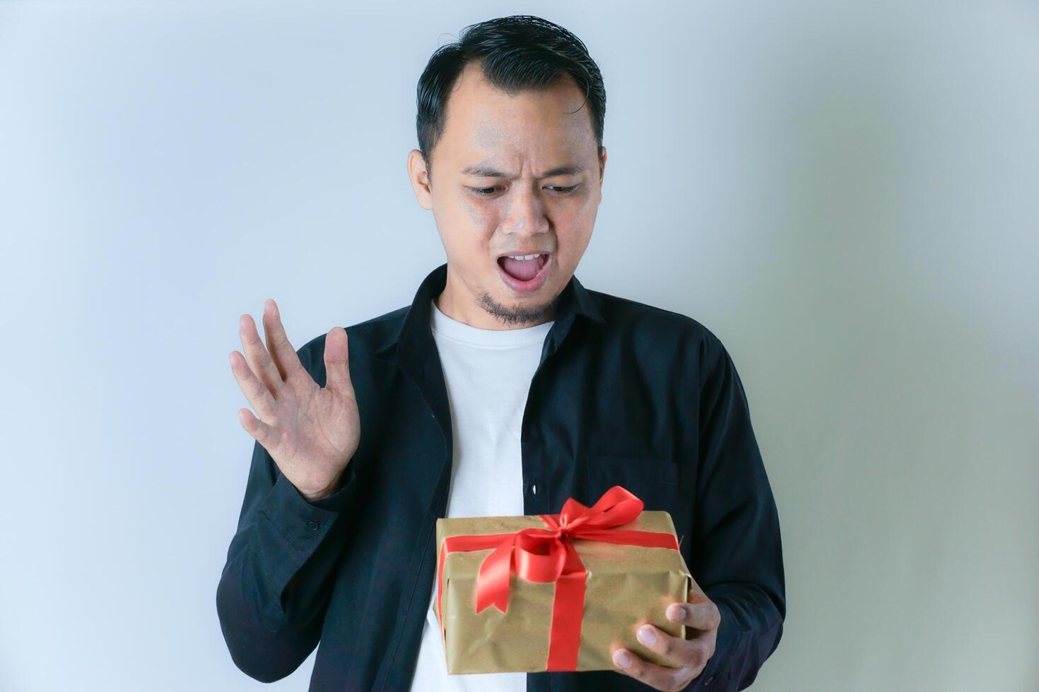 A man in a black shirt and white t-shirt was surprised while holding a gold gift box with a red ribbon, against a white background. photo