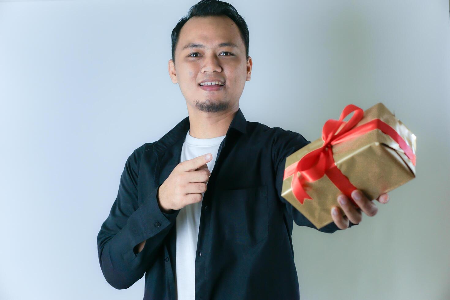 A man wearing a black shirt and a white T-shirt is smiling and pointing to a gold gift box wrapped with a red ribbon, against a white background photo
