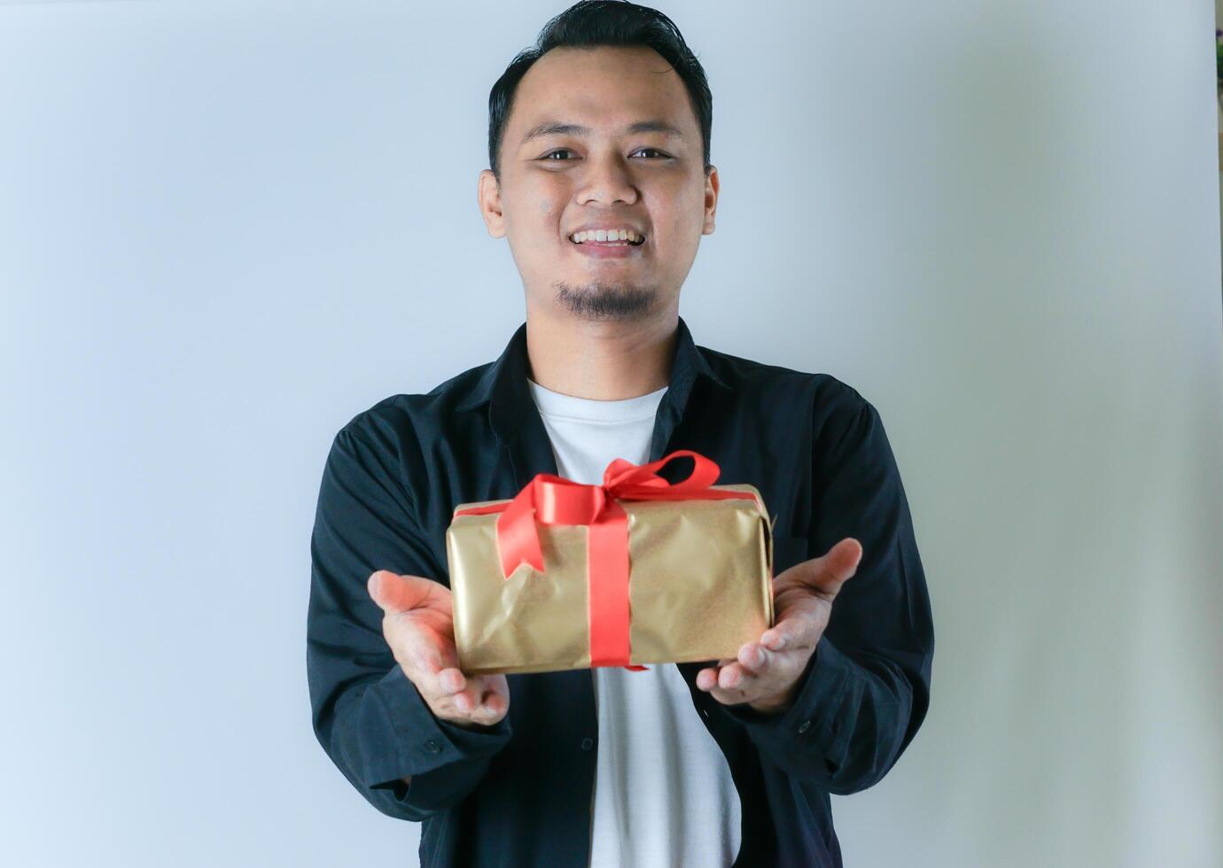 A man wearing a black shirt and a white T-shirt is smiling while presenting a gold gift box wrapped with a red ribbon, against a white background. photo