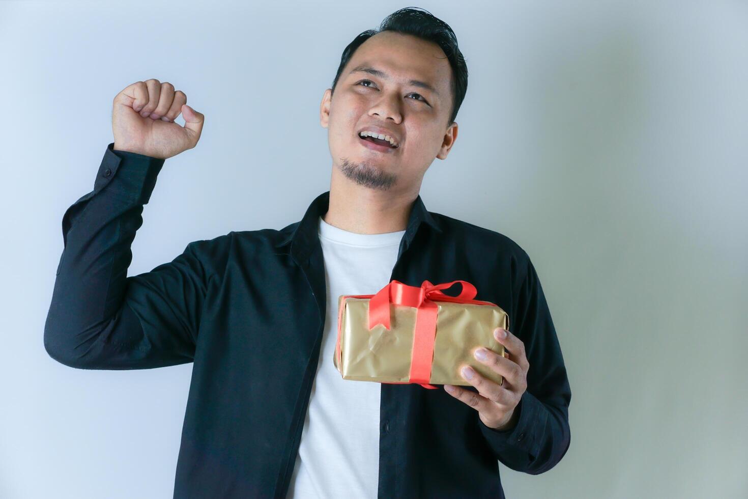 A man in a black shirt rejoices while holding a gift wrapped with a red ribbon, showing excitement and appreciation. photo