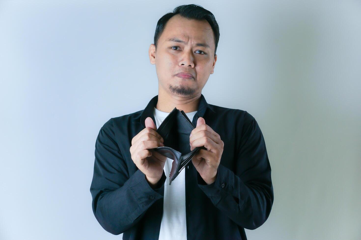 A man in a black shirt and white t-shirt shows an empty wallet with a worried expression, conveying financial difficulties or lack of money. photo