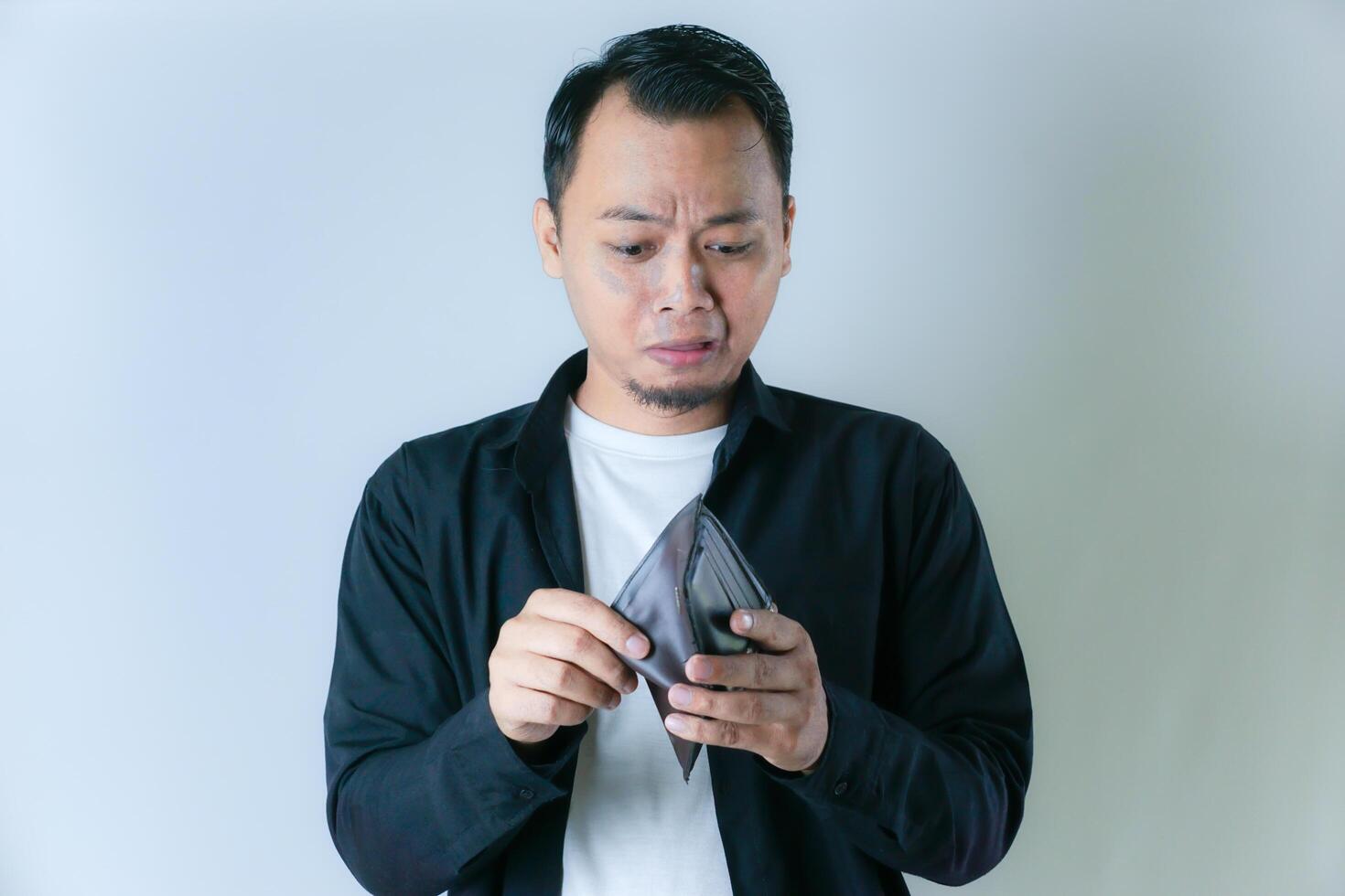 A man in a black shirt and white t-shirt shows an empty wallet with a worried expression, conveying financial difficulties or lack of money. photo