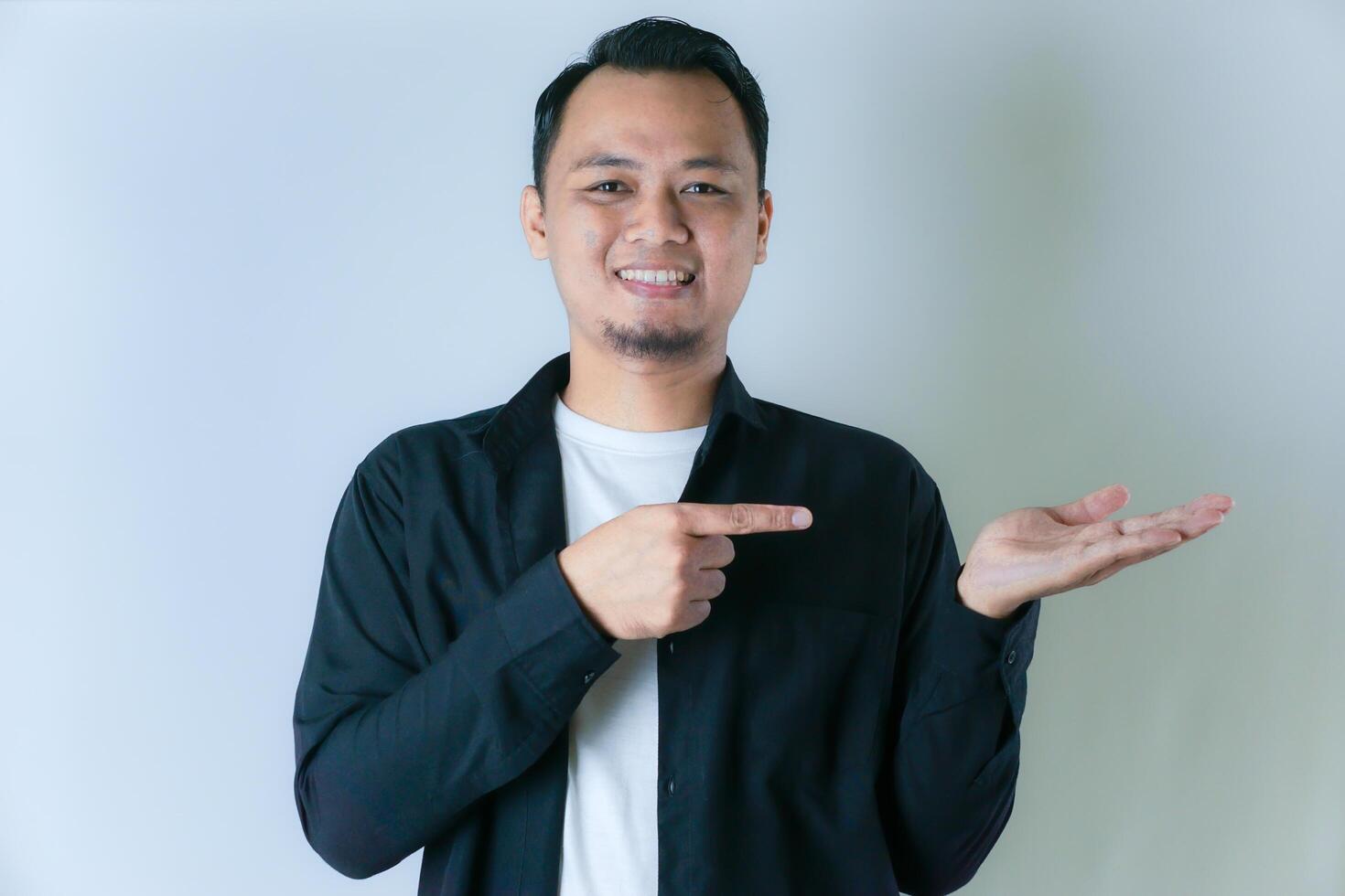 A smiling man in a black shirt and white undershirt is pointing to his open palm, as if presenting or showcasing something against a plain background. photo