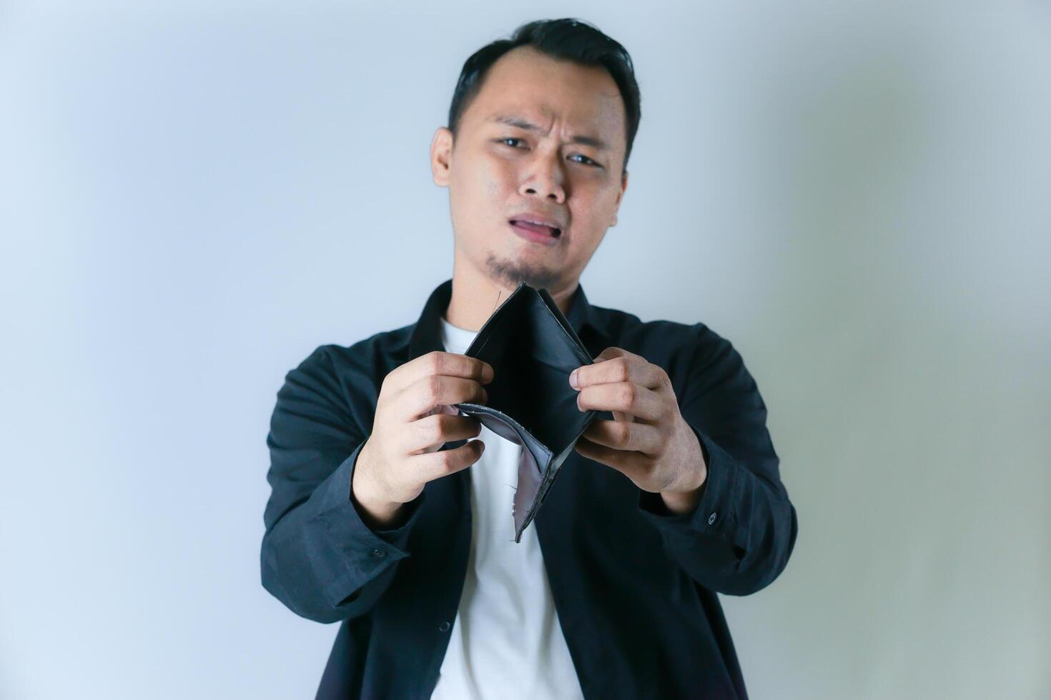 A man in a black shirt and white t-shirt shows an empty wallet with a worried expression, conveying financial difficulties or lack of money. photo