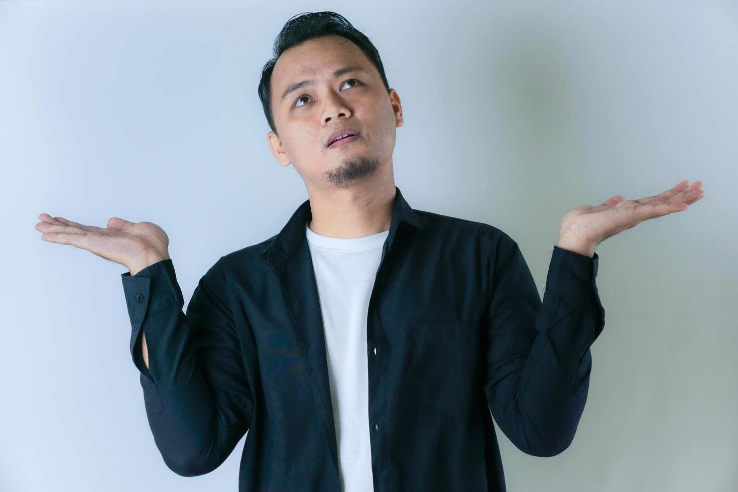 A man in a black shirt and white t-shirt looks confused, raising both hands with a questioning expression against a plain white background. photo
