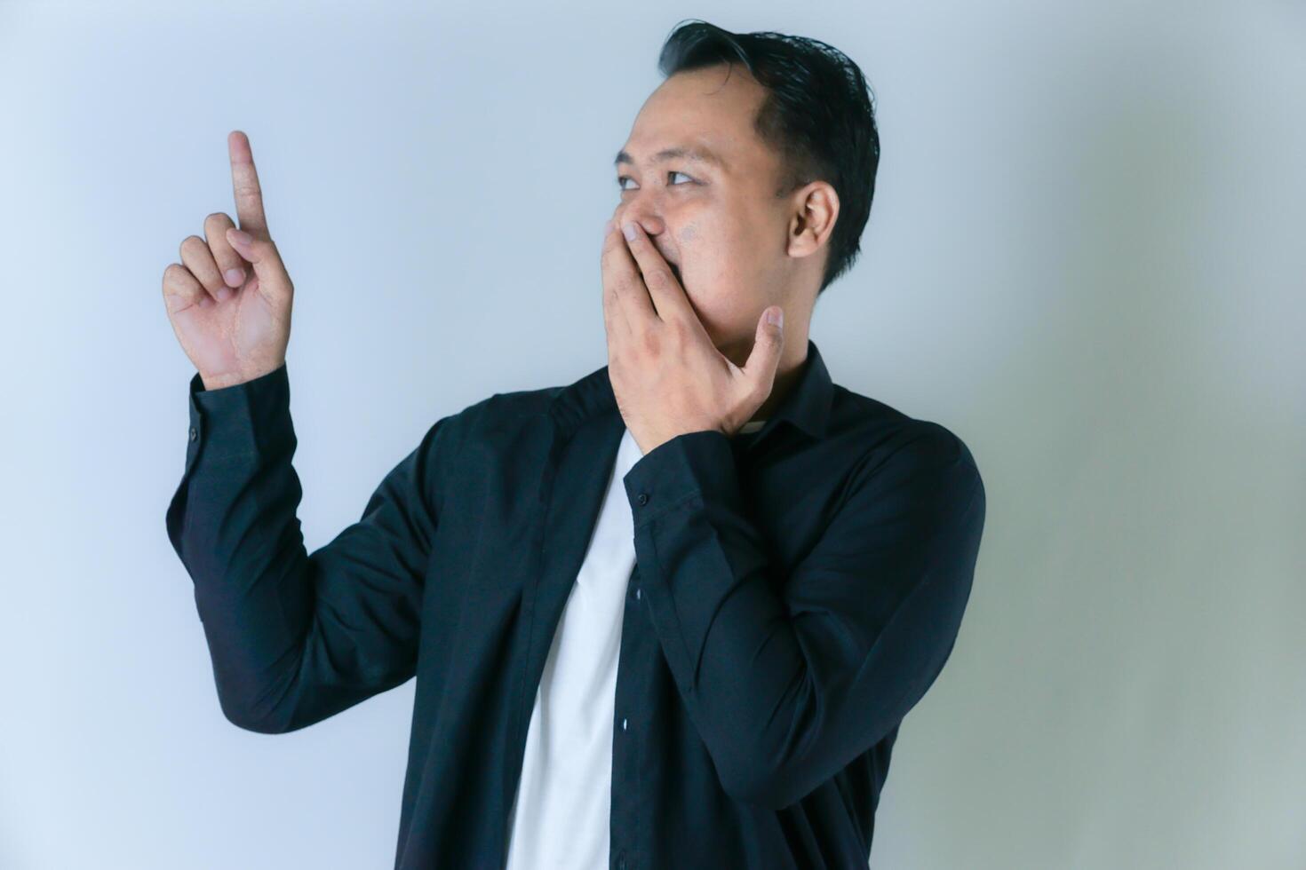A man in a black shirt and white t-shirt holds his hand to his mouth and points upwards thoughtfully, standing against a plain white background. photo