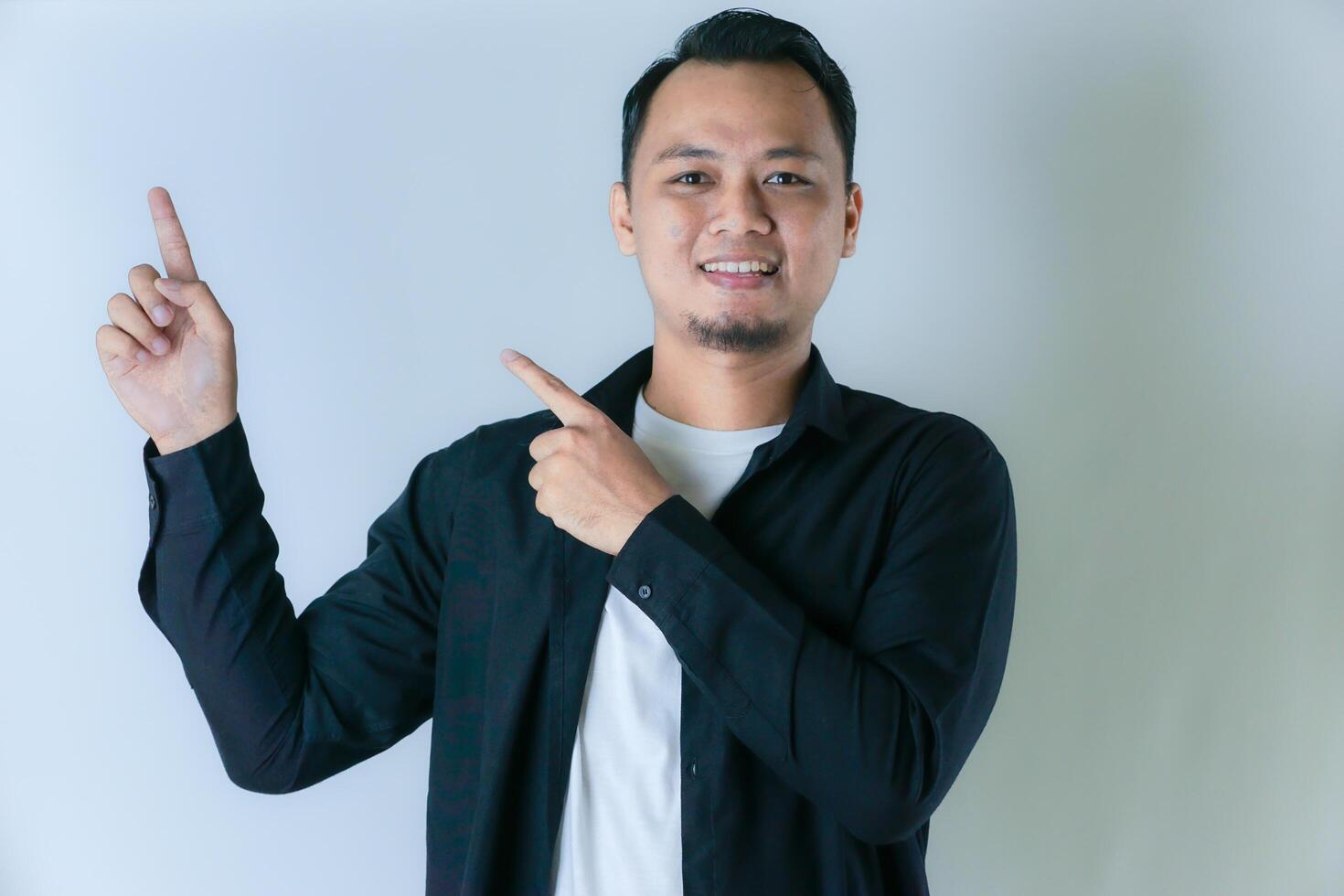 A man in a black shirt and white t-shirt smiling in a pose while pointing upwards with his two index fingers, on a white background photo