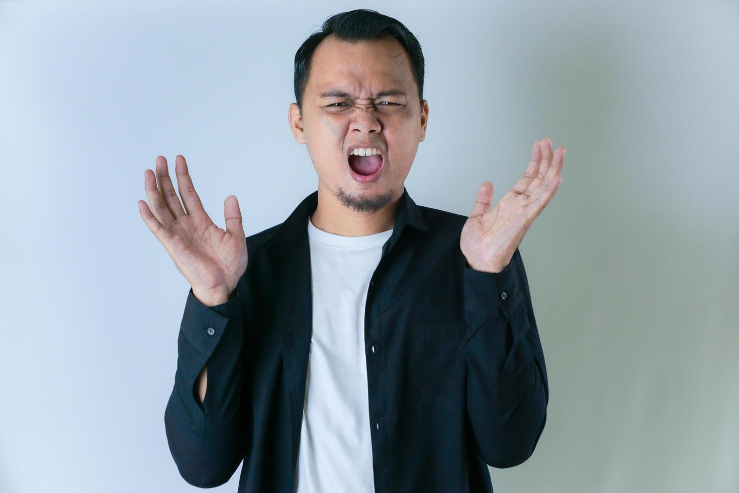 A man in a black shirt and white t-shirt is expressing frustration or anger, with his hands raised and a tense facial expression against a plain background. photo