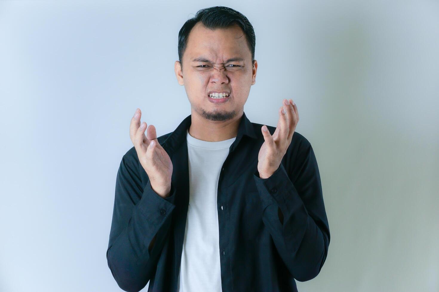 A man in a black shirt and white t-shirt is expressing frustration or anger, with his hands raised and a tense facial expression against a plain background. photo