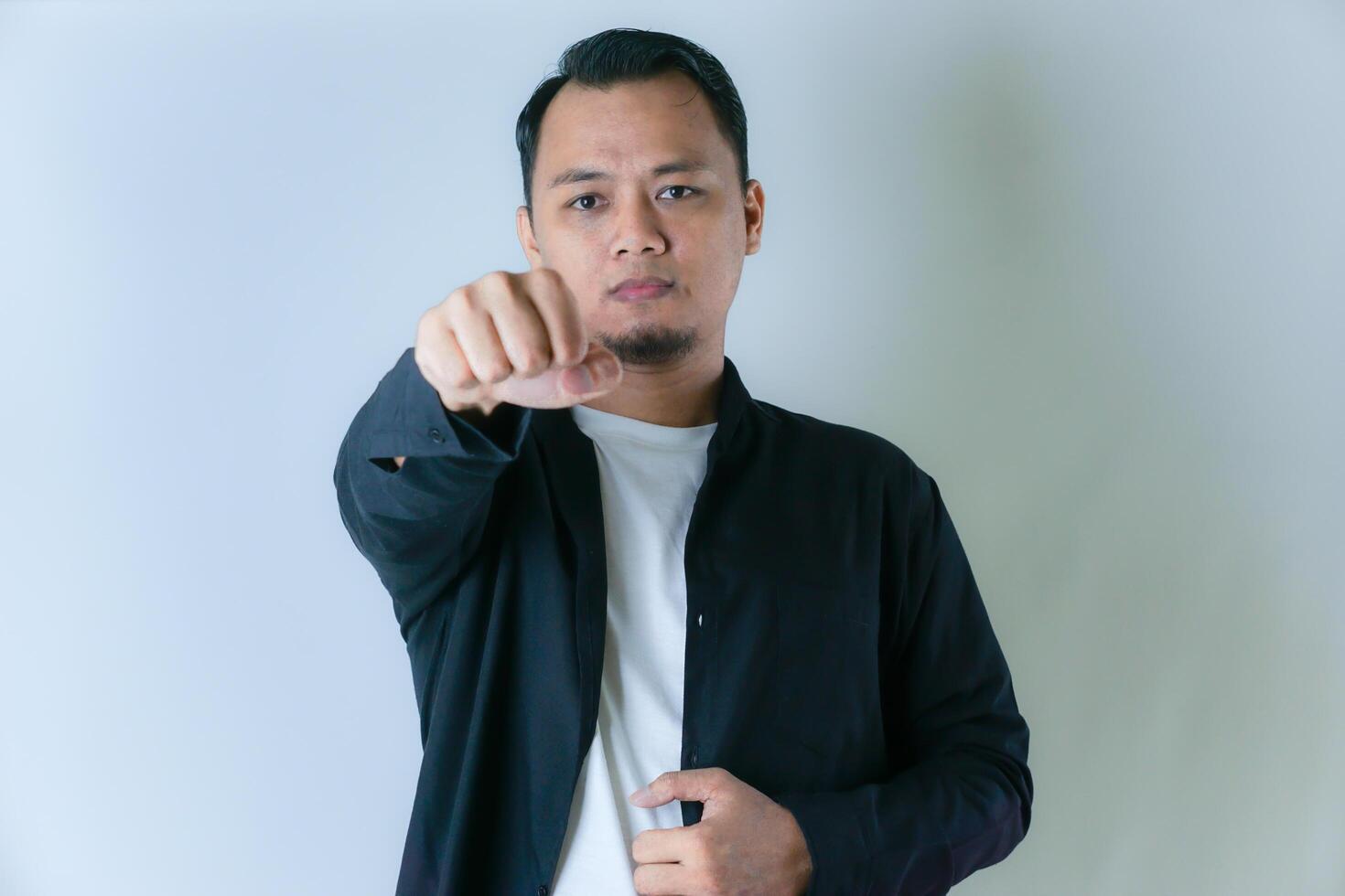 A man in a black shirt and white t-shirt is confidently pointing forward into the camera, exuding determination and strength against a plain background. photo