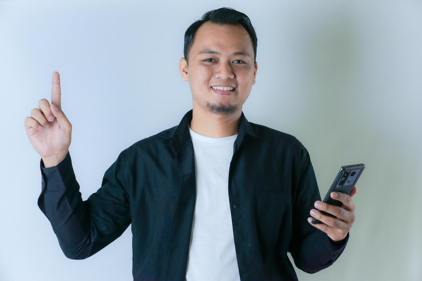 A smiling Asian man in a black shirt and white t-shirt holds a cell phone while pointing upwards photo