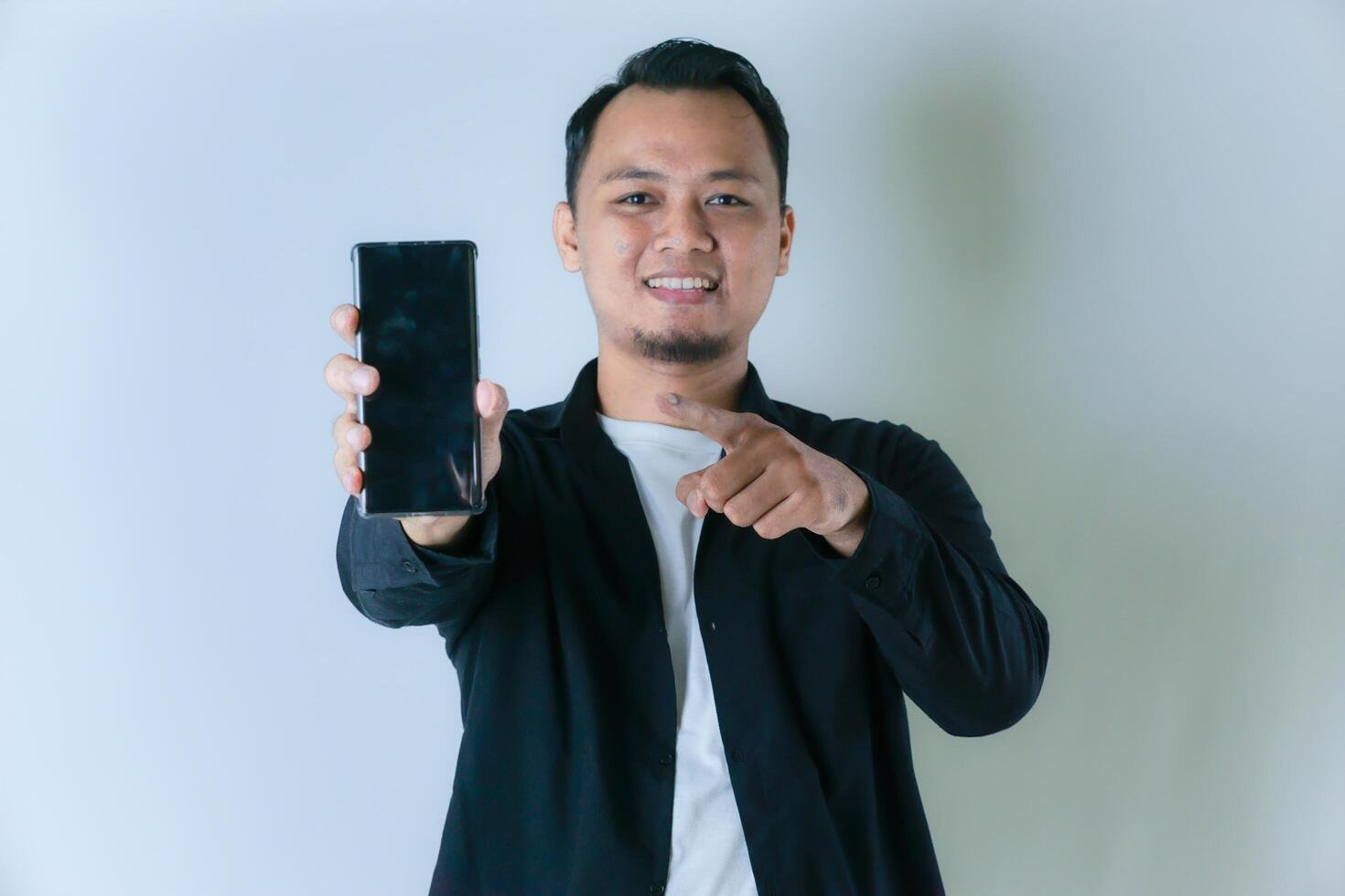 A smiling Asian man in a black shirt and white T-shirt is holding a cell phone and pointing at it. photo