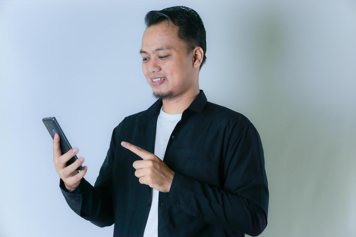 A smiling Asian man in a black shirt and white T-shirt is holding a cell phone and pointing at it. photo