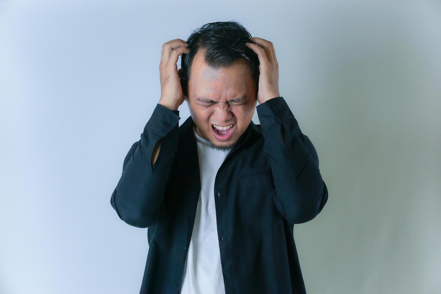 A man in a black shirt and white t-shirt is expressing frustration or anger, with his hands raised and a tense facial expression against a plain background. photo