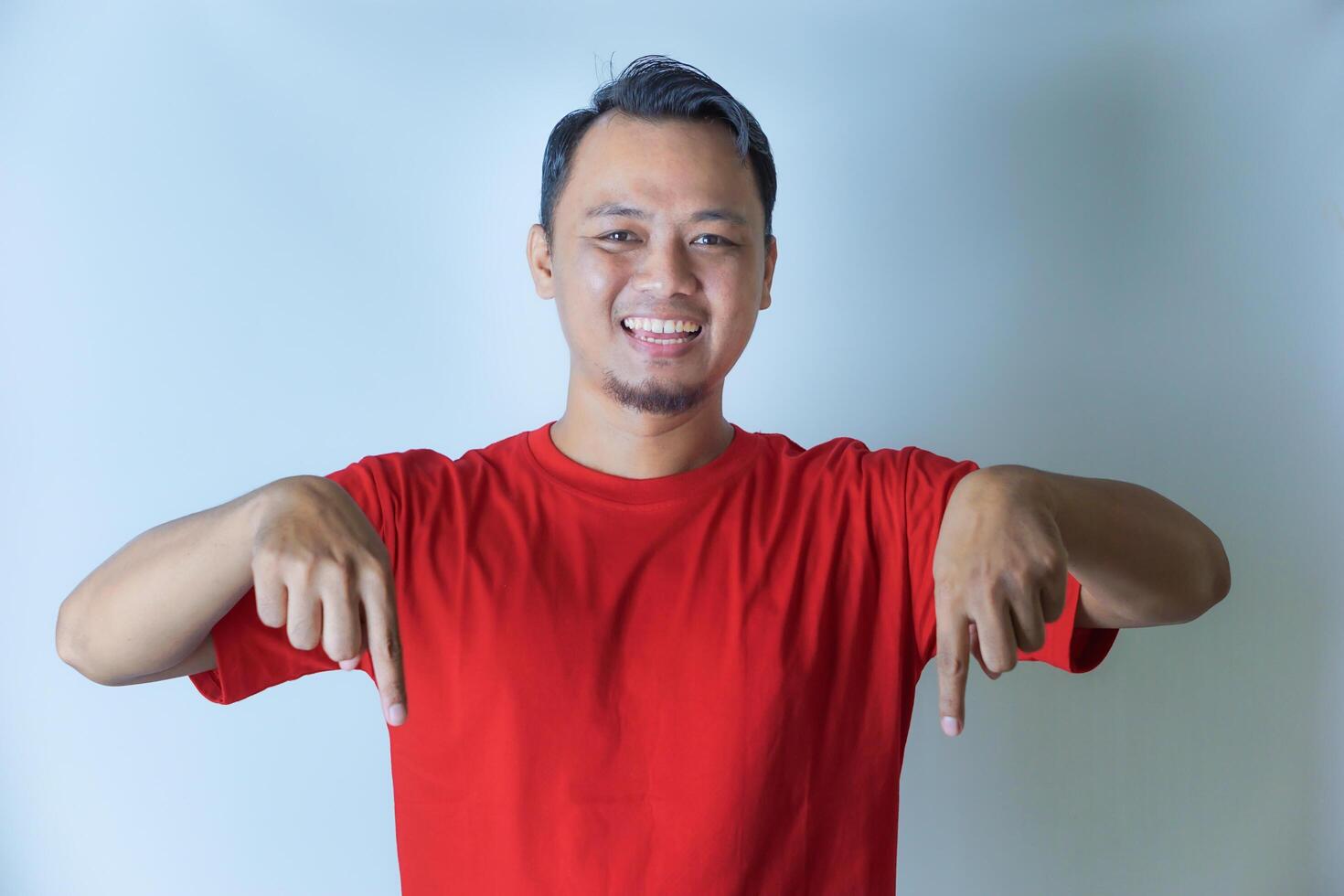 A man in a red shirt is smiling and pointing down with both index fingers, looking cheerful and excited. isolated on white background photo