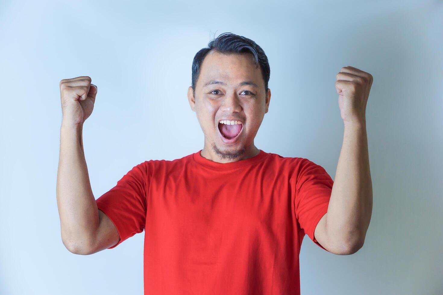 A man in a red shirt joyfully raises his fists in a celebratory gesture, smiling widely against a plain white background. photo