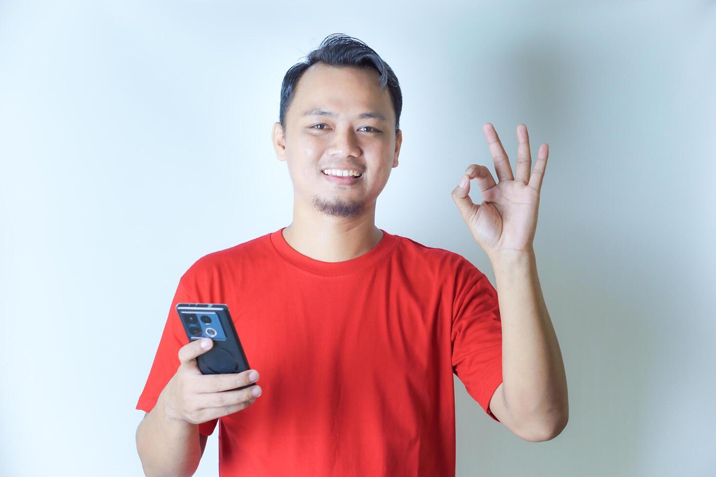 A man in a red shirt holds a smartphone in one hand, giving an OK hand gesture with a smile, indicating satisfaction or approval. photo