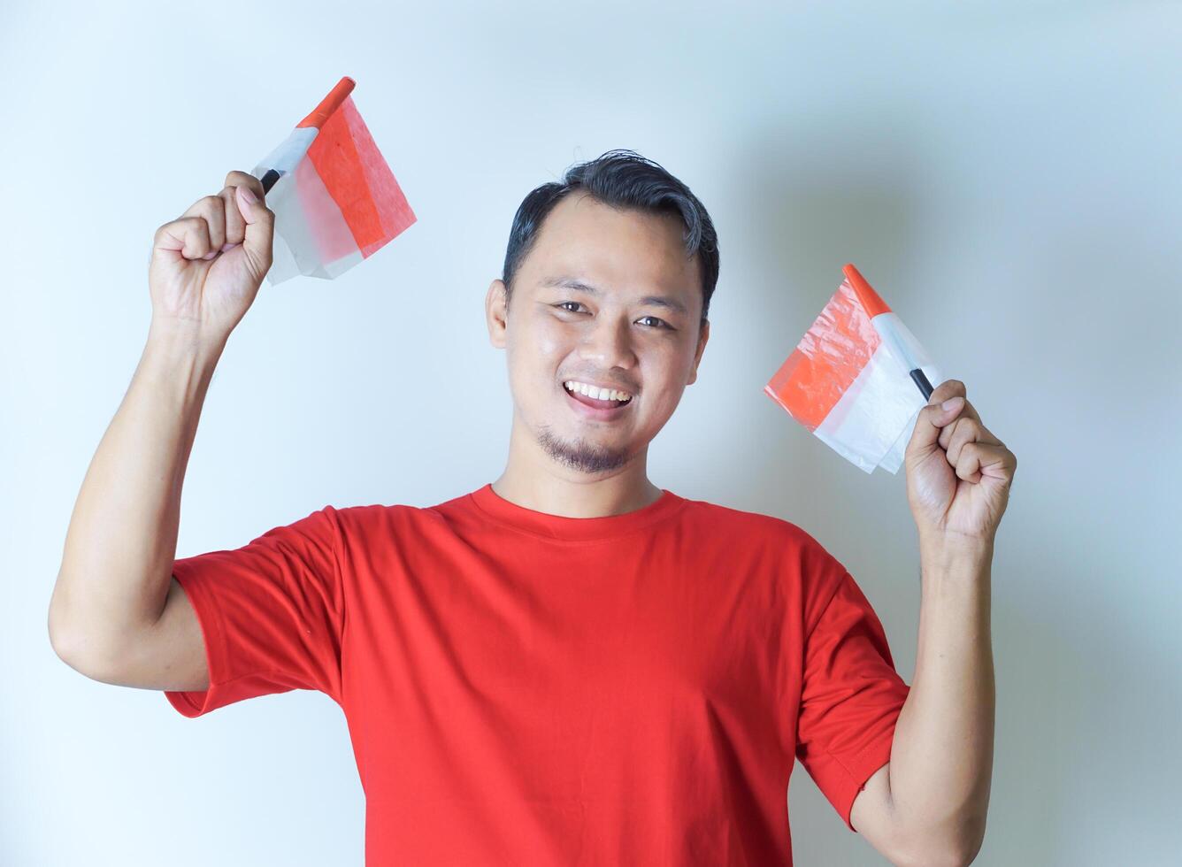 A man in a red shirt smiles while holding two small Indonesian flags, the concept of celebrating Indonesia's independence day photo