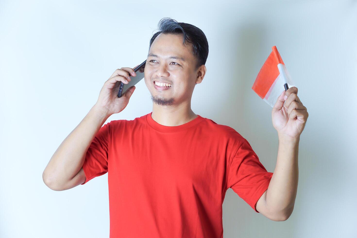 A man in a red shirt smiles while holding the Indonesian flag and talking on his phone, embracing the concept of celebrating Indonesia's independence day. photo