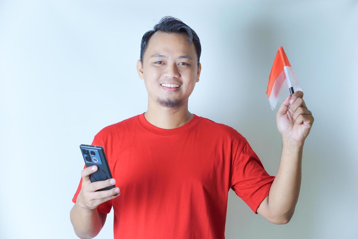 A man wearing a red shirt holds a smartphone and waves an Indonesian flag, showcasing his pride in the concept of celebrating Indonesia's Independence Day. photo