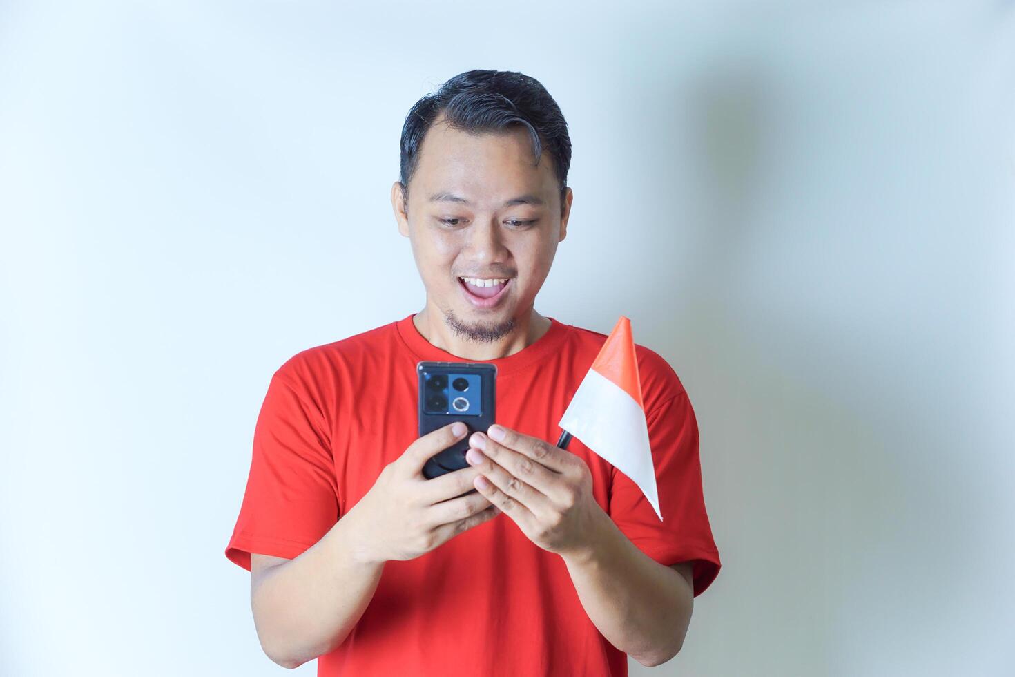 A man wearing a red shirt is joyfully looking at his phone while holding an Indonesian flag, the concept of celebrating Indonesia's independence day. photo
