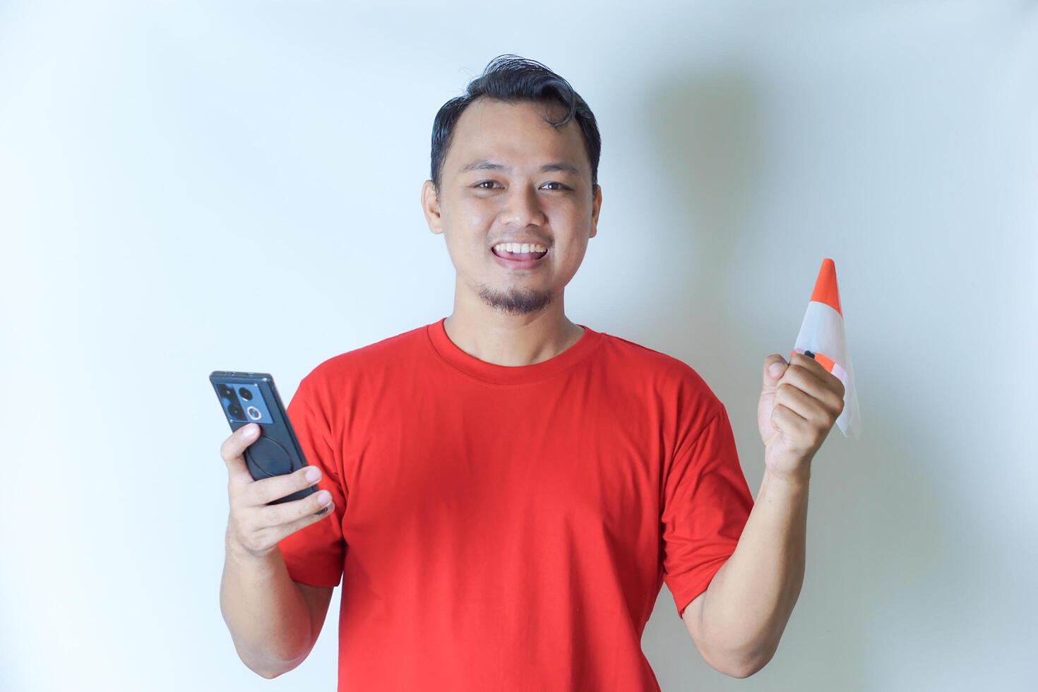 A man wearing a red shirt holds a smartphone and waves an Indonesian flag, showcasing his pride in the concept of celebrating Indonesia's Independence Day. photo