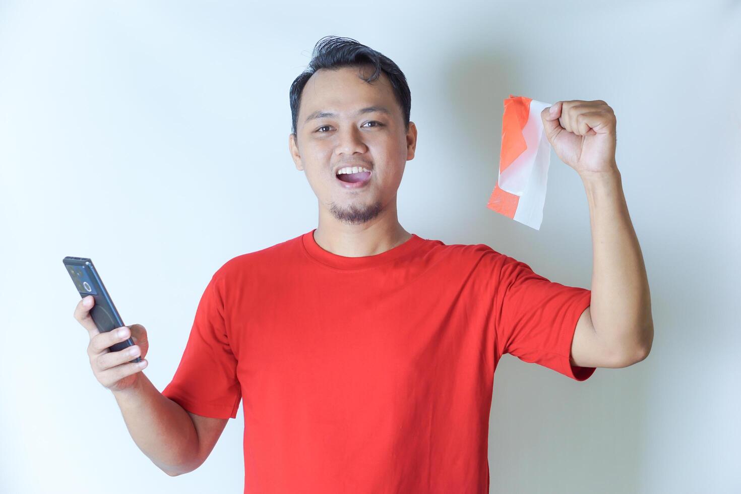 A man wearing a red shirt holds a smartphone and waves an Indonesian flag, showcasing his pride in the concept of celebrating Indonesia's Independence Day. photo