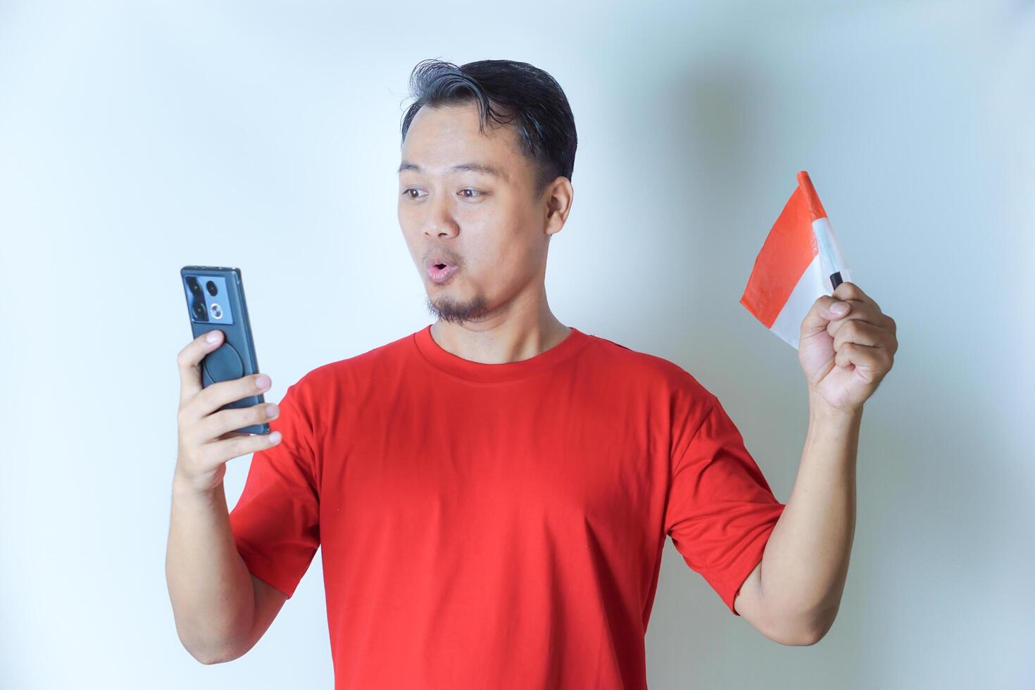 A man in a red shirt holds an Indonesian flag while looking at his phone with excitement, embodying the concept of celebrating Indonesia's independence day. photo