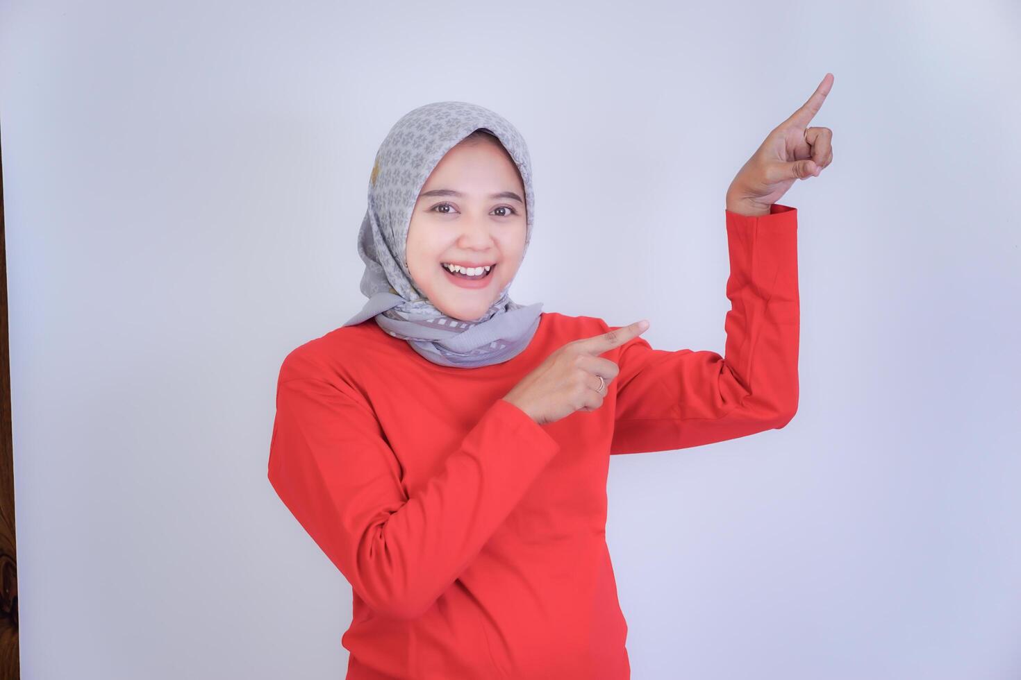 A woman in a red shirt and hijab smiles and points both fingers to the upper right, highlighting something. photo