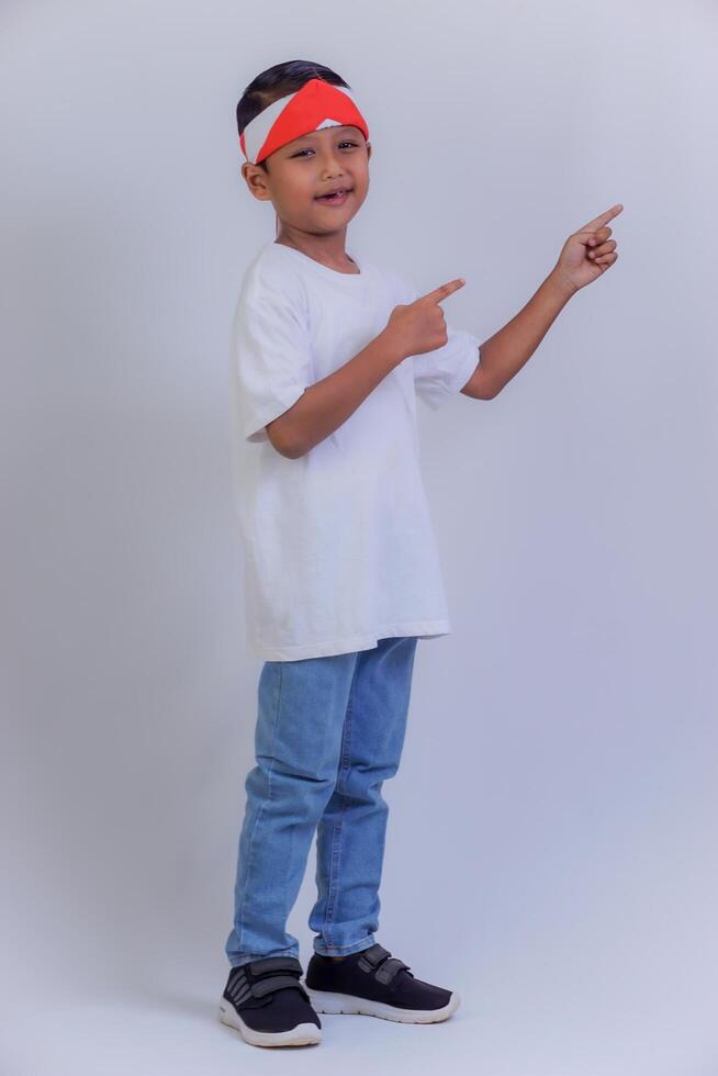 A boy pointing to the side wearing a white shirt, jeans, and a red and white headband, smiling confidently. photo